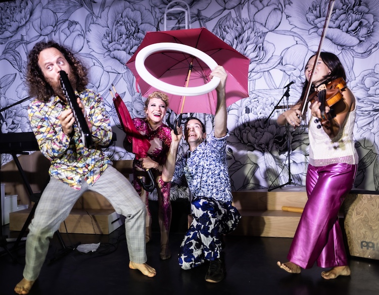 against a black and white floral backdrop, a quartet of performers happily pose with their instruments and props