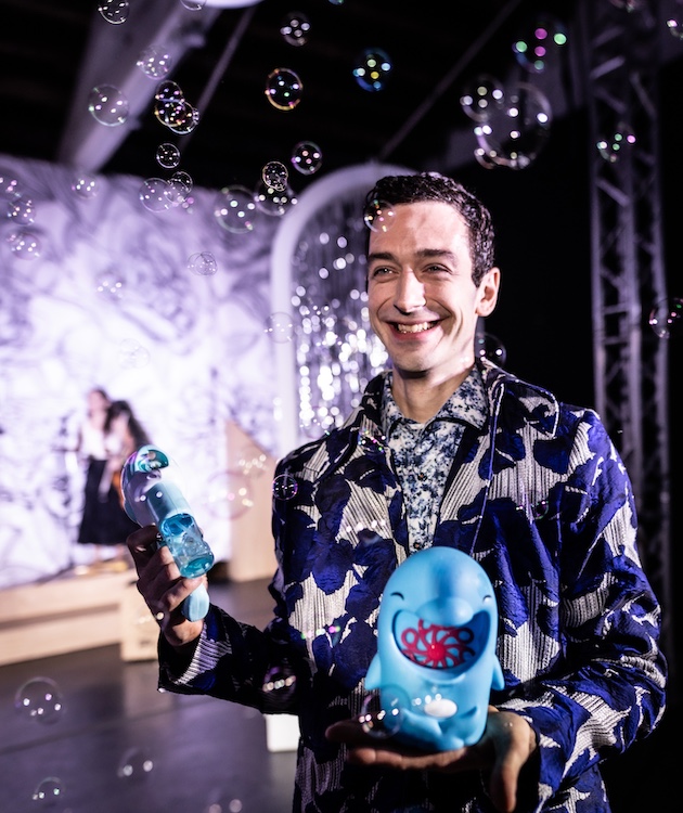 kyle driggs in the foreground wearing a  colorful navy patterned suit surrounded by bubbles blowing from a hand-held bubble blower. He is all smiles. 