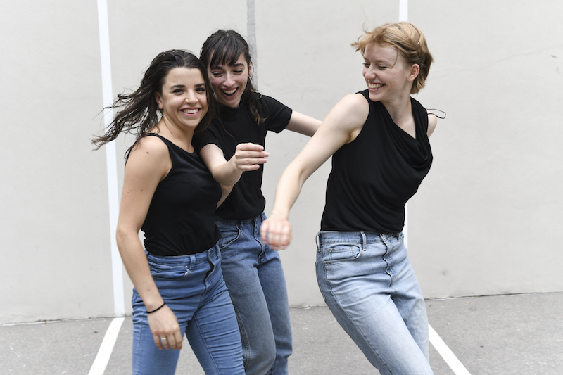 Three women smile wearing black tanks and jeans.