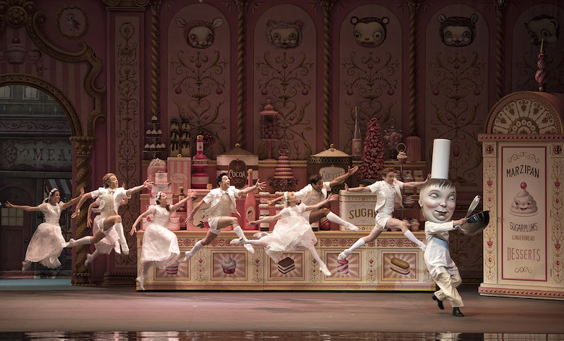 A group of young dancers dressed in white leap buoyantly in the air. A dancer in a larger-than-life chef masks teases them with sugary confections.