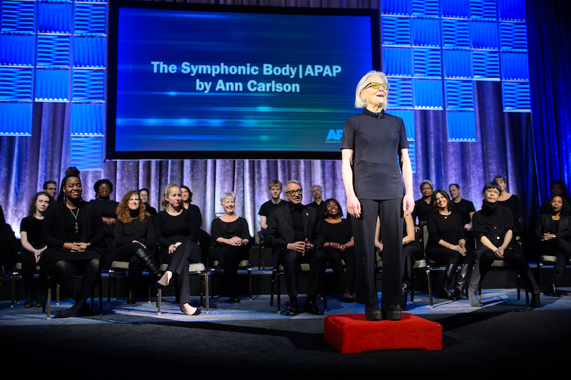 Ann Carlson, all in black, stands in front of her symphonic body chorus who are seated and also wearing black.