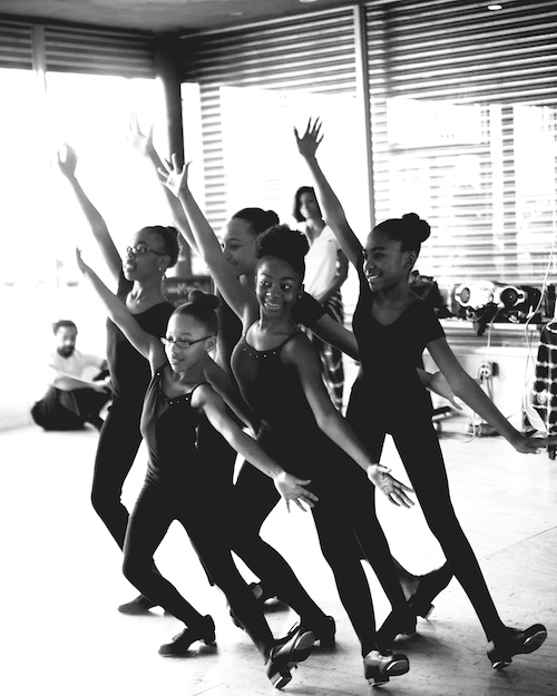 A group of young students in black tights, leos and tap shoes. They pose in a tight grouping with on foot extended out. Their arms splayed in a diagonal.