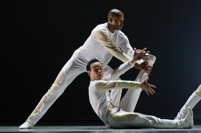 Two dancers in white. One sits on the floor with his legs straight and scissor-like. The other is in a wide stand behind him. Leaning towards his right bent leg. 