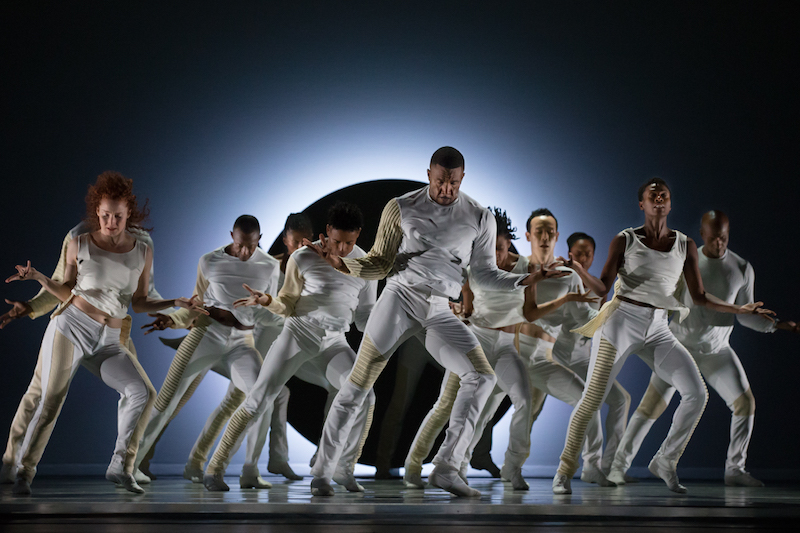 An ensemble of dancers all in white stand with their arms stretched out to their sides. Heads are down.