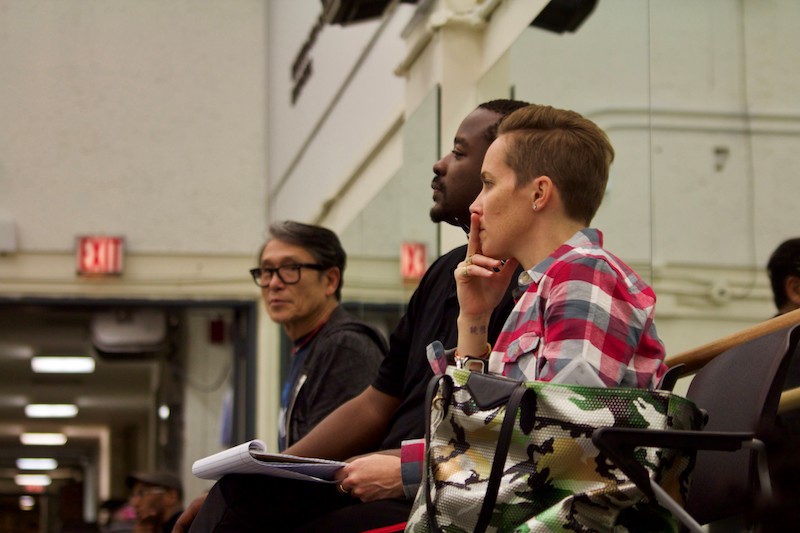 Three Ailey creatives sit against the studio mirror as they watch rehearsal. Elisa Clark sits with a notebook in hand.