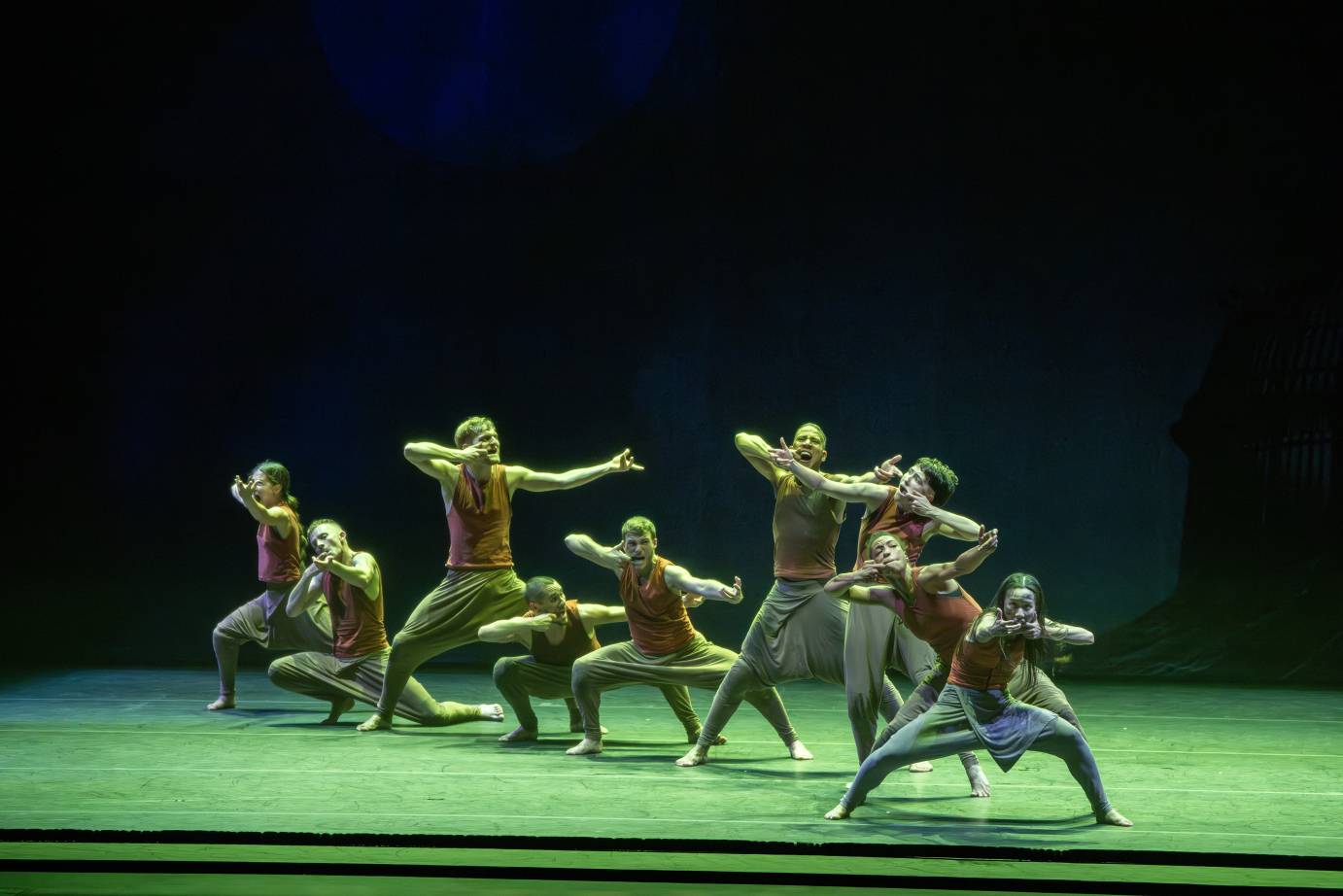 under a green light, a group of dancers similarly dressed in sleevless orange shirt sand grey pants stand in a varied array of positions. All of them see to be aiming rifles, hunting. 