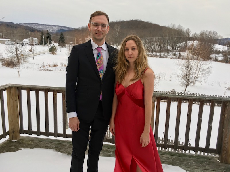 Neal Medlyn in a suit and Maggie Cloud in a red silk evening gown pose for a photo outside where there is snow and trees.