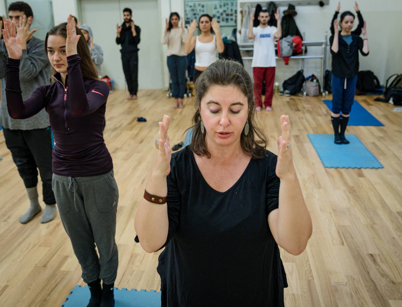 Anabella Lenzu with fair skin brown hair wearing dangling blue earrings and a black outfit leads a class of students. All are standing on blue yoga mats with eyes closed, legs together and arms bent at the elbow, hands framing the face.