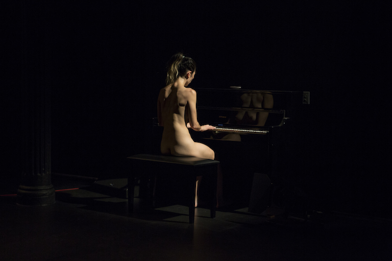 Angela Goh sits naked at a piano and plays the instrument. Her back is to the audience. 
