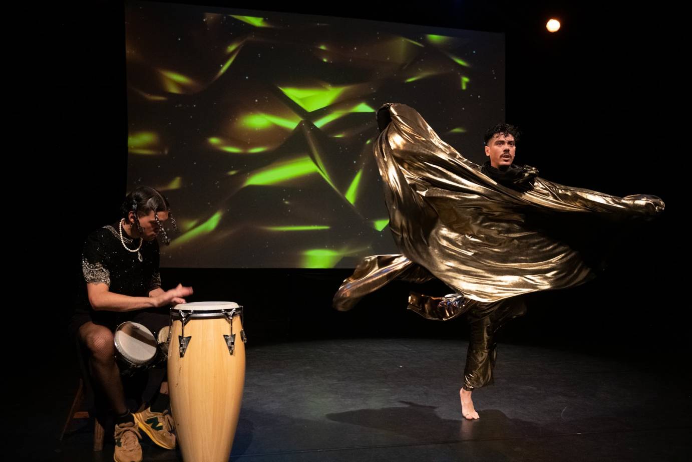 A drummer dressed in black shirt and shorts and textured stockings plays the conga while a mustachioed man dances in swirling gold fabric. Projected behind them is an undulating gold backdrop pierced by bright green glints..