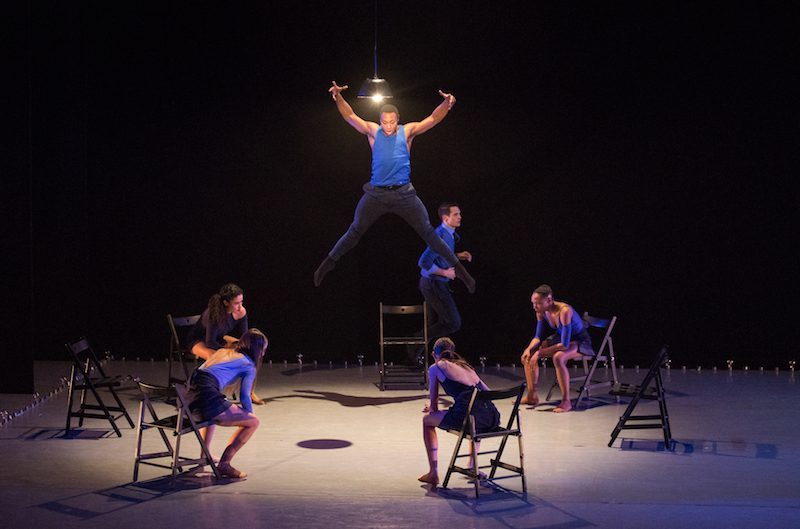 A man in a blue shirt and black slacks leaps high into the air as dancers sit in chairs in encircling the artist