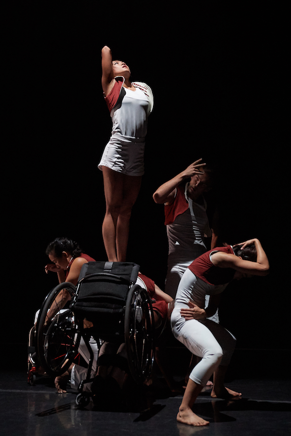 Dancers in white and red create a tall tableau. A few dancers contort around another performer in a wheelchair. One dancer stands above them.