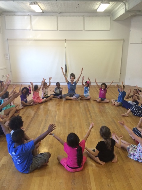 A group of kids sit in a circle with arms raised above their heads. Dance artist teacher Donna Costello leads them in the exercise.