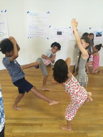 A group of kids play. One has his leg in the air. Two children have their palms together with their arms raised above their head. Another stands on one leg.