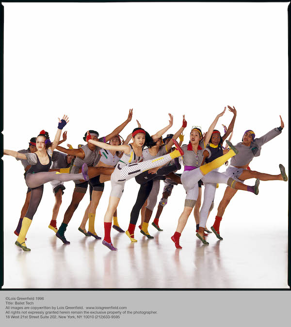 A group of Ballet Tech students in colorful leotards, tights, shoes and leggings, lift their right leg and flex their foot. They look out at the camera 