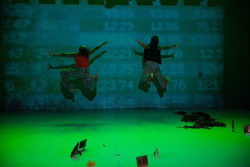 Two women, with their back facing the audience jump into the air. The stage is in lit in green hues. Numbers are subtlety projected onto the back wall.