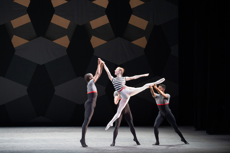 against geometric shape in tan and black a ballerina in a striped top and white shirt skirt jumps in arabesque held by two male dancers wearing grey and black unitards. One man holds her uplifted arm, the other, her highly extended leg. The tone is joyful