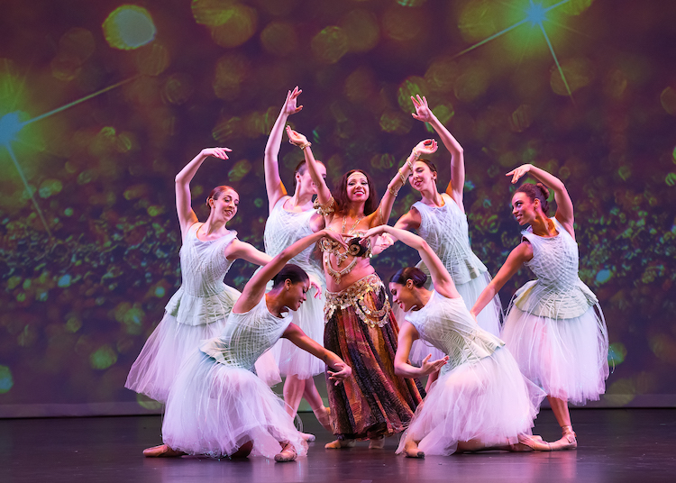 a belly dancer poses amongst a group of ballet dancers in white tutus
