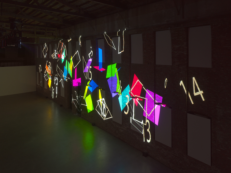 a projection of colored numbers of various sizes jumbled on a long brick wall at Pioneer Works