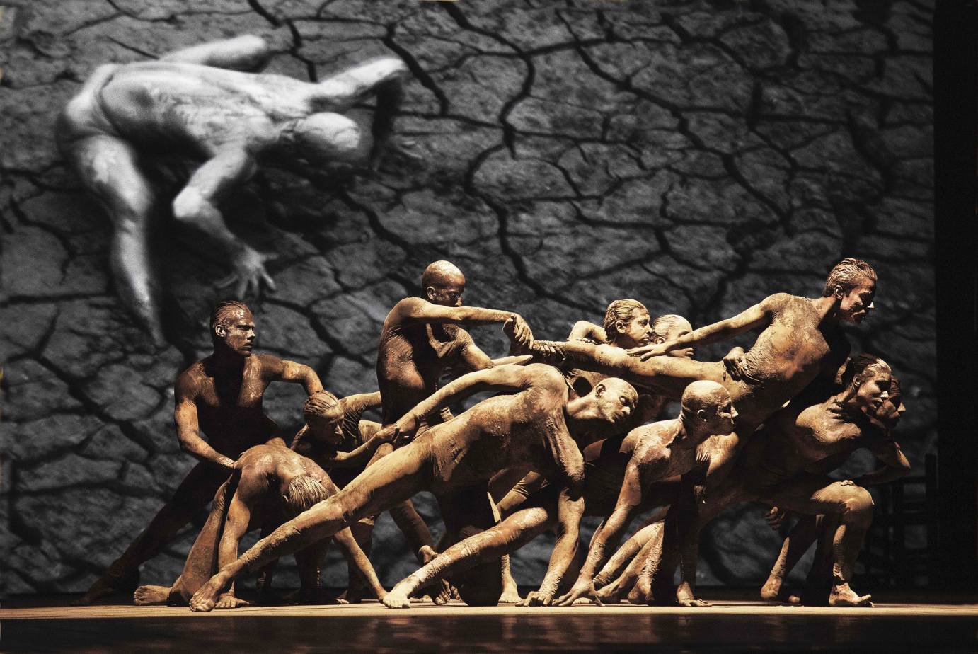 Against a black and white film, mud-caked dancers lunge in a clump