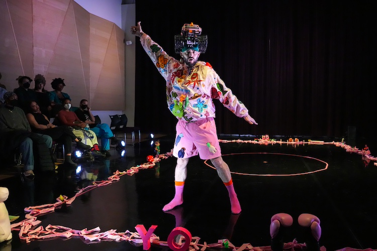 a colorful room defined by a large circle of pink ribbons and toys that holds a smaller pink circle,sets the stage for Christopher's ritual to lost childhood. He wears pink: a windbreaker, shorts, socks, and a pink toy monster truck on his head, the outfit decorated with colorful toys scattered on his costume as if thrown on.