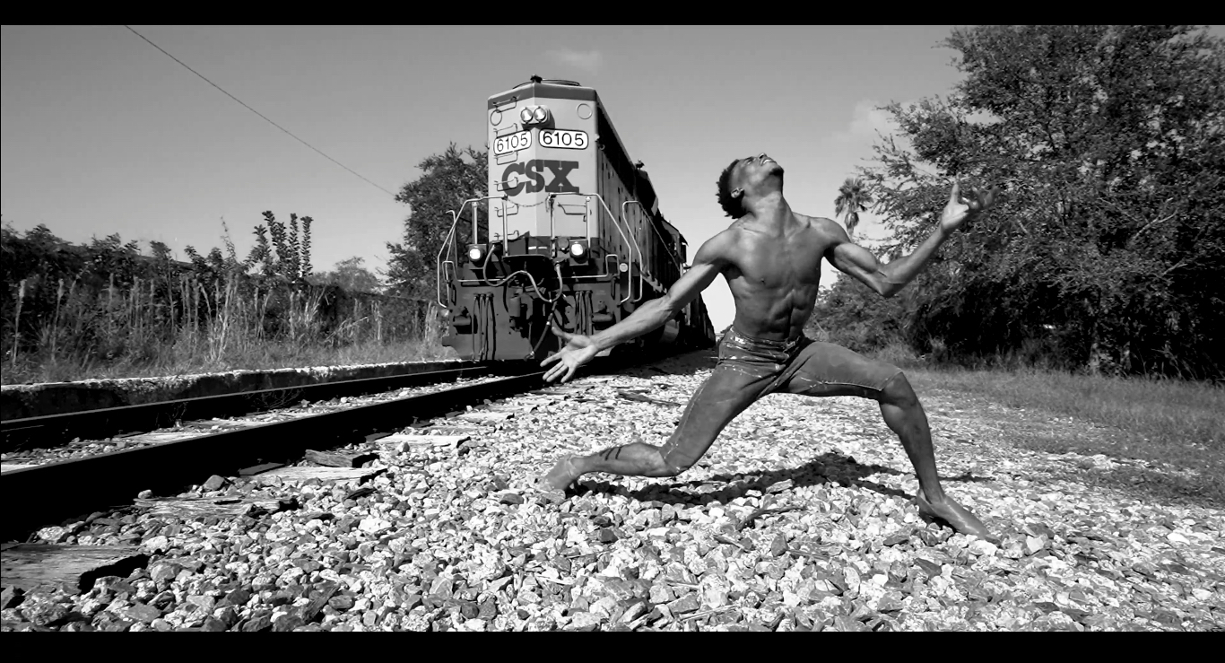 Beside a train track, Jared Brunson lowers into a kneel, his arms extended, his fingers splayed as a train approaches