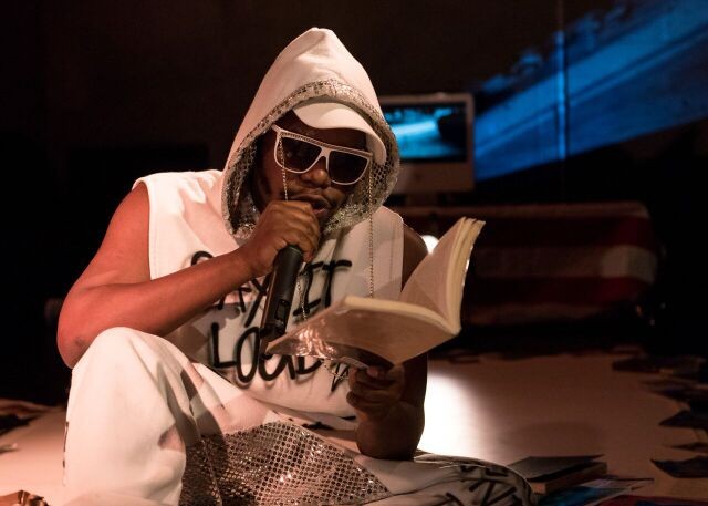 Jaamil Olawale Kosoko wearing a white hoodie and sunglasses while reciting poetry from a book.