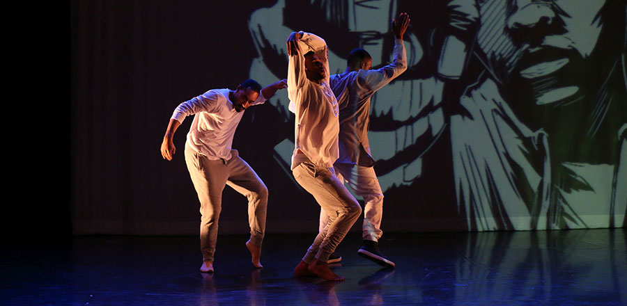 Three African-American dancers dressed in loose smocks and trousers on stage.