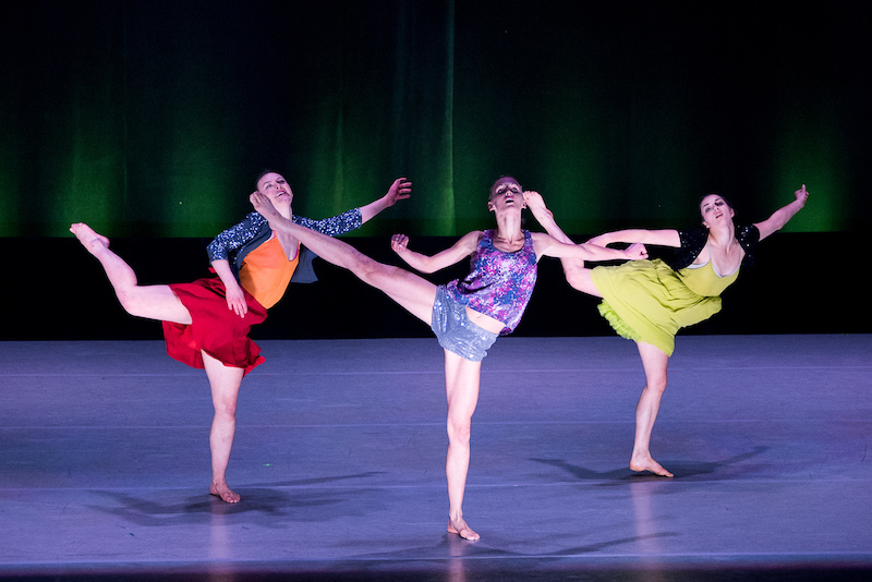 Three women in colorful blouses and skirts lift one of their legs in turned in and contorted positions