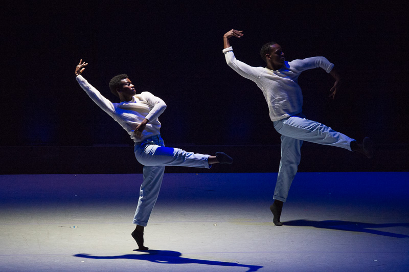 Two dancers in white pants and shirts relve on one leg while their other leg is extended into an air. They slightly arch their backs.