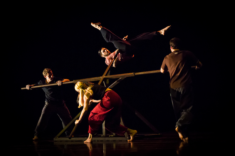 Four dancers surround a geometrically structured wood object. One dancer is in midst of hoisting himself up onto one of the structure's spindles. 
