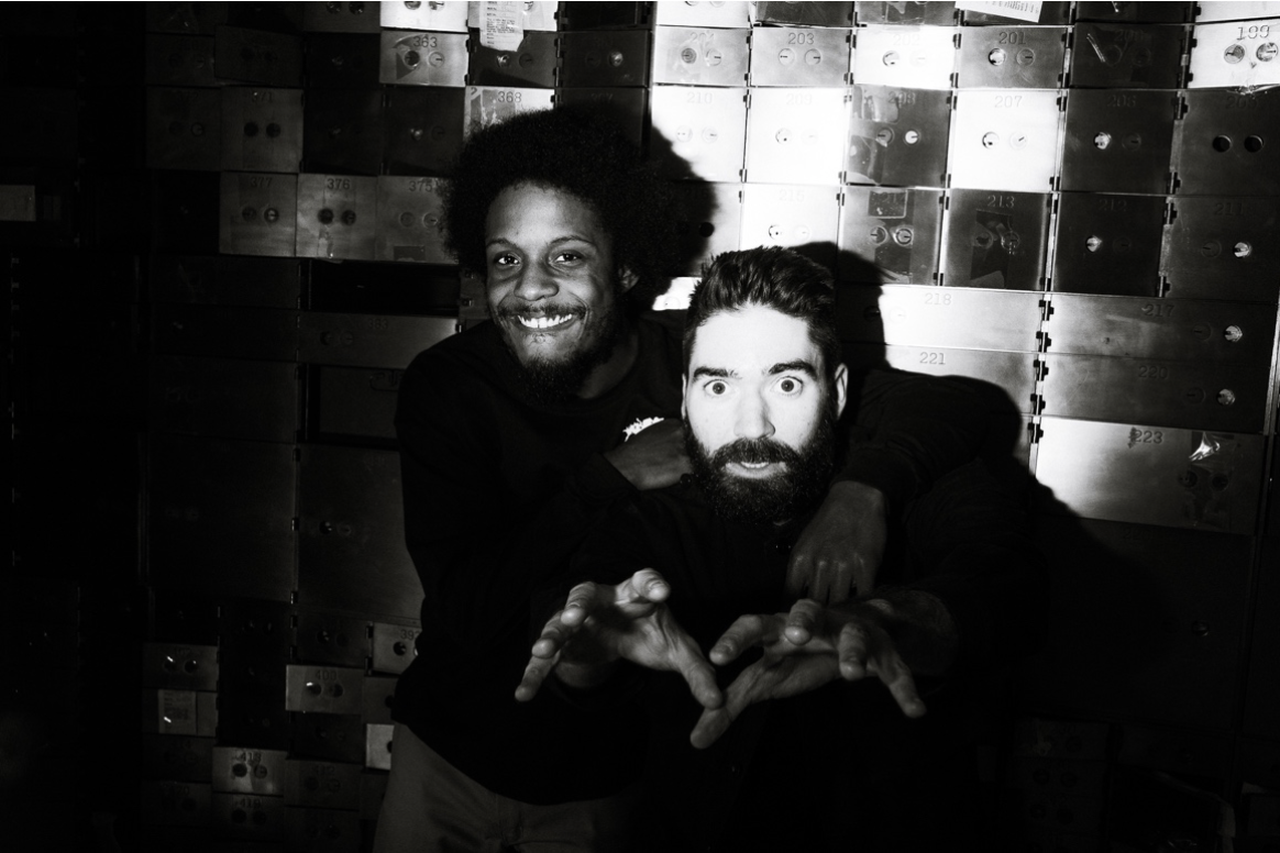 A black and white photo of two men in front of stacked boxes. One man with short Afro and mustache is smiling, the other sports a wide-eyed expression and clawed hands that dramatically reach toward the camera.