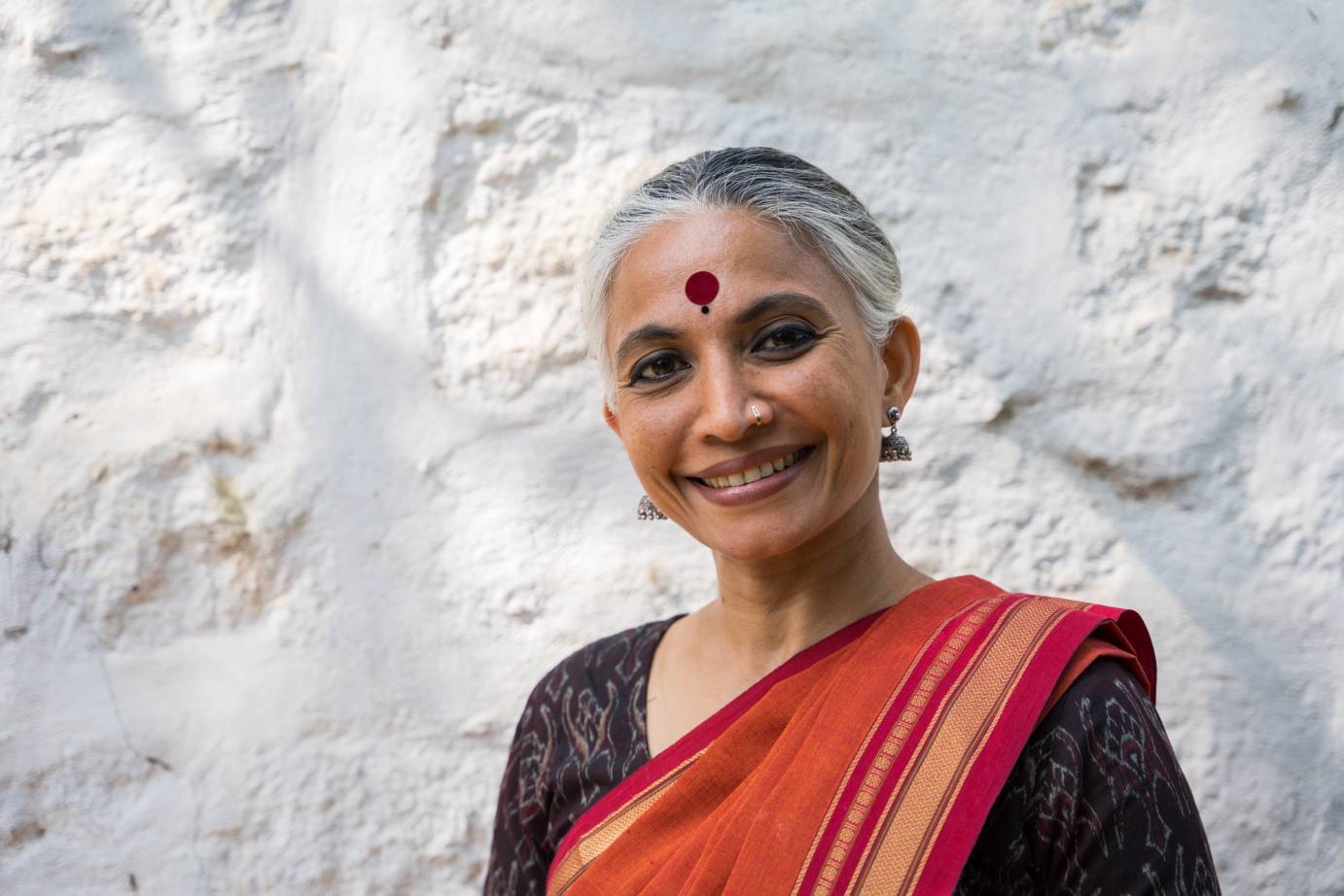 A grey-haired smiling woman, with a deep red bindi (dot) in the middle of her forehead, dangling earrings and nose ring, wears a long red and yellow striped fabric over her left shoulder beneath which she wears a dark and white patterned top.