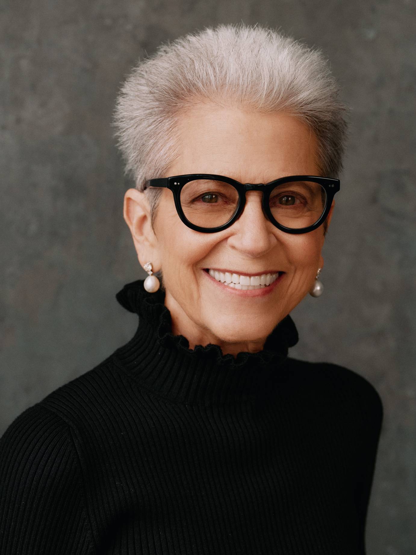 A smiling woman, with short-spiked white hair, big black framed glasses, dangling pearl earrings, and ruffled black turtleneck looks out to the camera.