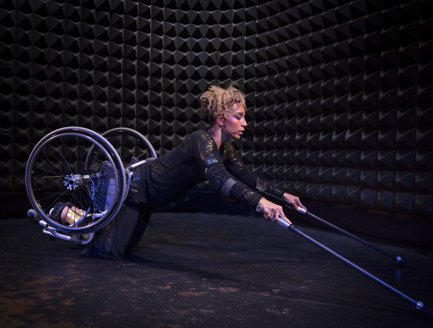 In a wheelchair, a woman balances on her knees. Her arms are extended, and she holds two sticks. 