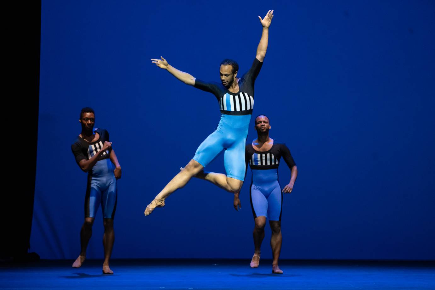 three men with dark skin dance in unitards with blue bottms and white and black striped tops... as two men in the background appear to be jogging , one man in the center jumps up in front of them