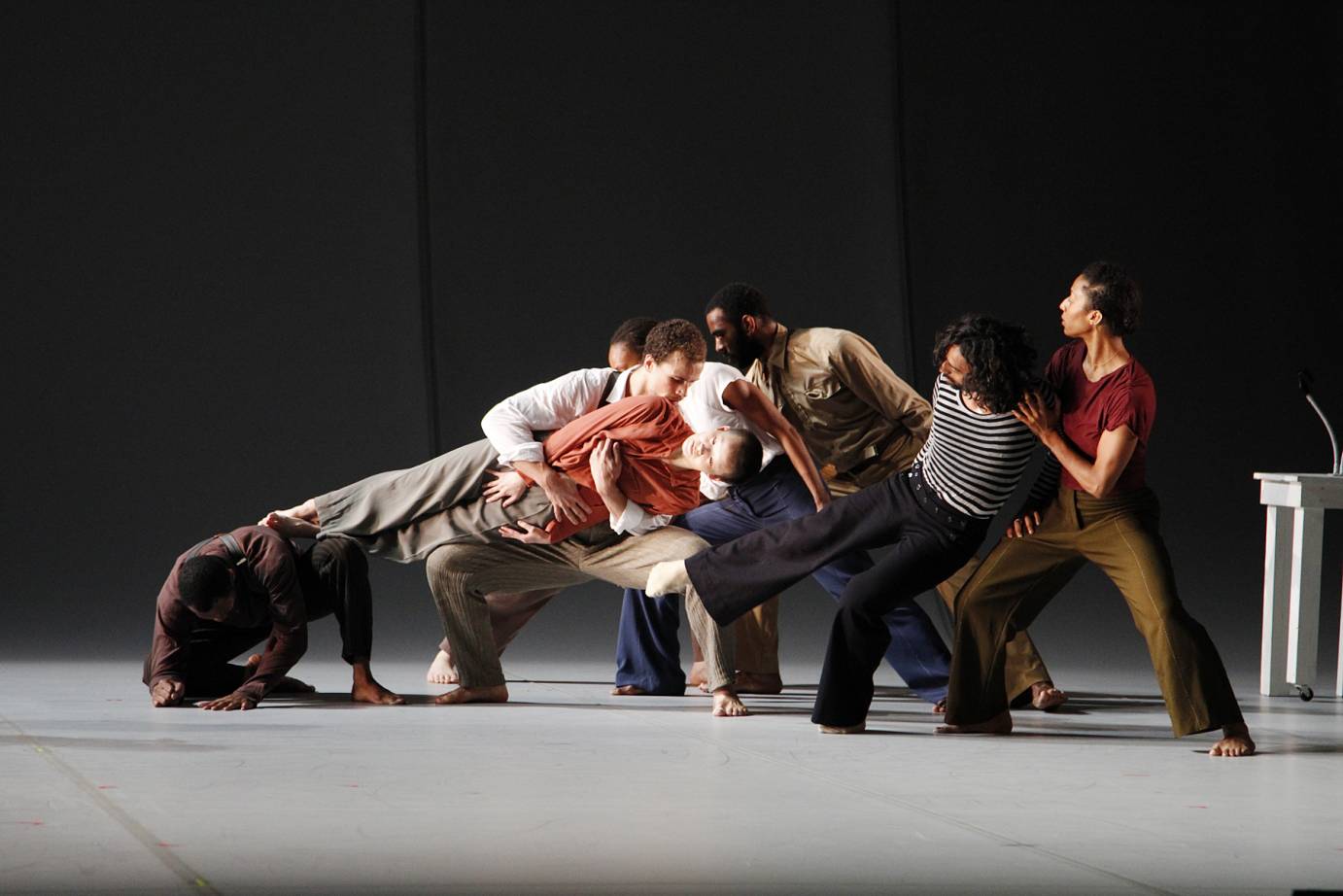 Dancers form a cascading chain with their bodies. They lean on one another. One dancer is held horizontally by the group.