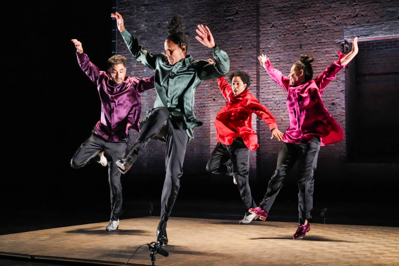 Four tappers jumping off the ground arms flung in the air dressed in purple, green, red, and magenta long-sleeved arms tossed in the air.