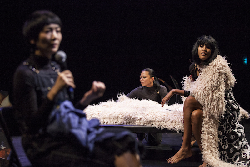 A woman in the foreground holds a mic up to her mouth. Another woman in a fur trimmed rope looks out to the audience. While another performer in the background smoothes a shag blanket that is atop a table.