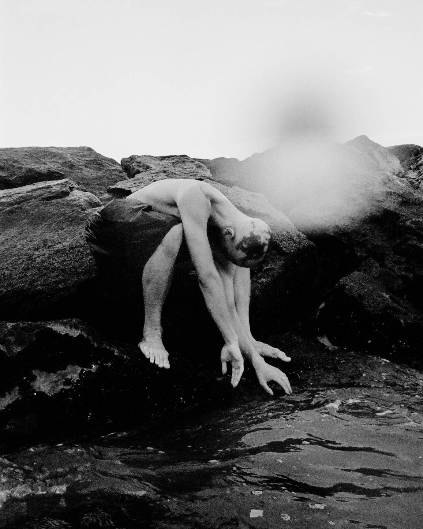 a black and white photo of a dancer crouched on boulders by the sea. The dancer wearing only pants or a skirt  melds into the landscape