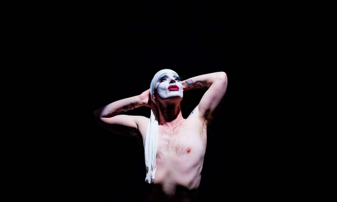 Dickie Beau, shirtless, places his tattoo-ed arm behind his head. His face, painted white with bright red lips, looks up to the stars. 