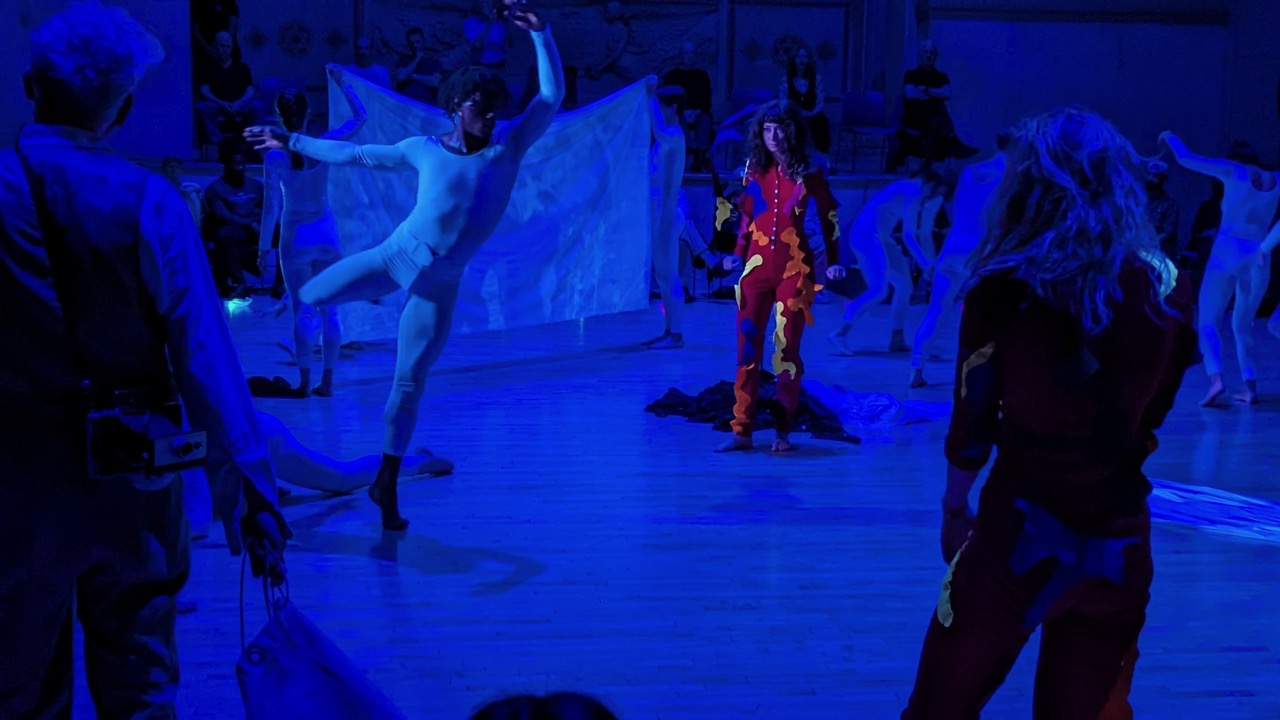 Male dancer in white unitard executing a back attitude. Woman in red costume next to him. Blue background with other dancers in white to the left of the dancers mentioned.