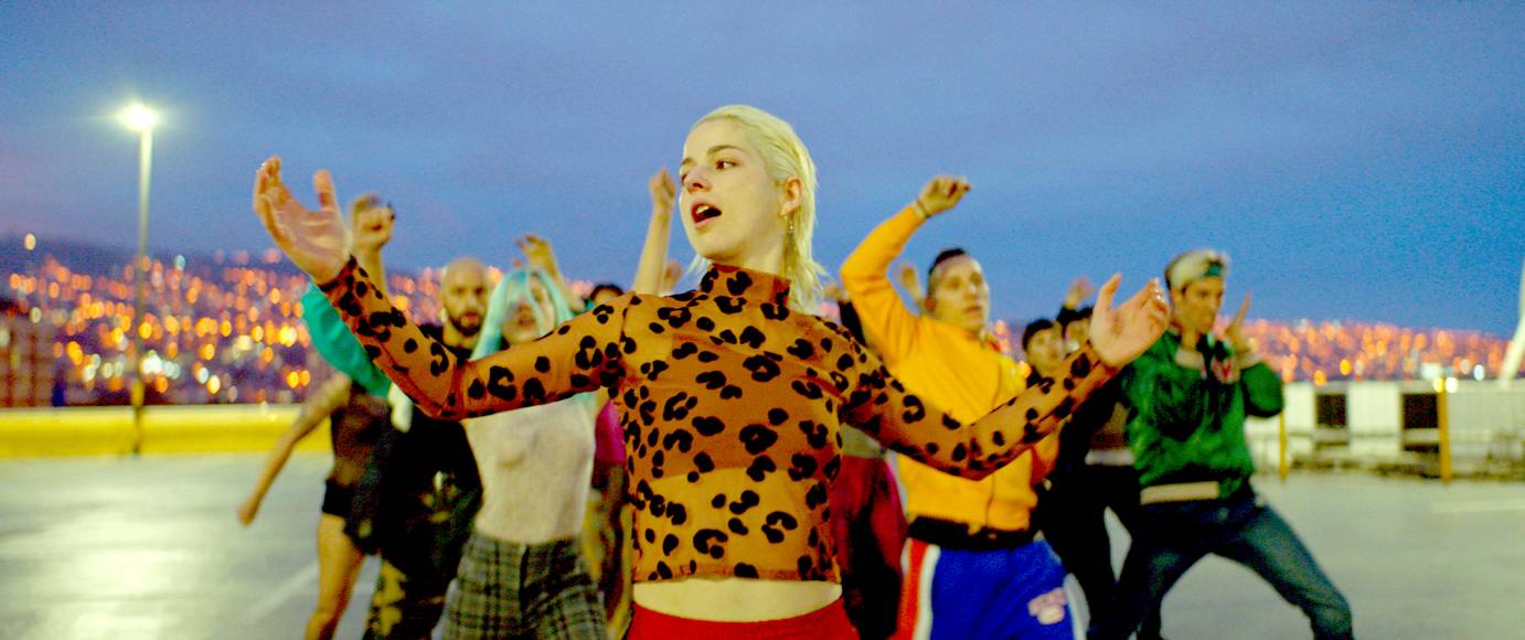 A woman in an animal print turtleneck dances in a parking lot with a group