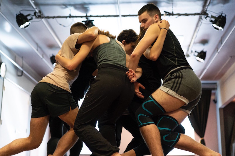 A group of dancers in rehearsal clothes huddle in a mass