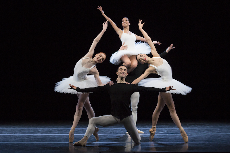 Dancers in white classical tutus pose in a tableau. One man is on his knee, two women are en pointe with the arms outstretched. And another male dancer lifts a woman onto his shoulder.