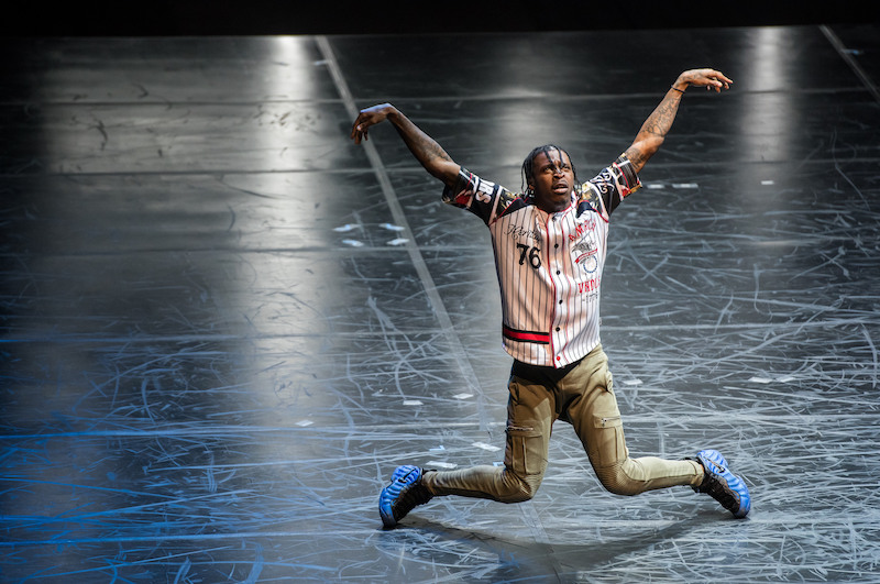 A FLEXN dancer in a baseball jersey looks like he's in mid-descent to the floor. His knees are turned inward and are parallel to the ground. His arms are outstretched and relaxed at the wrists as he being supported by an invisible bar. 