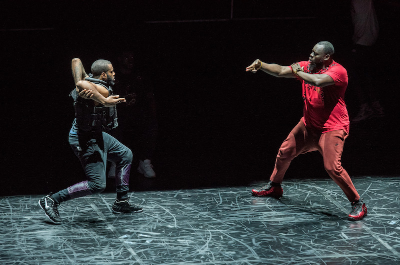 Two flexn dancers face one another. One has his elbows bent behind his upper back. His legs are in a crouched position. Another dancer, in all red, faces him, his right arm gestures to his counterpart.