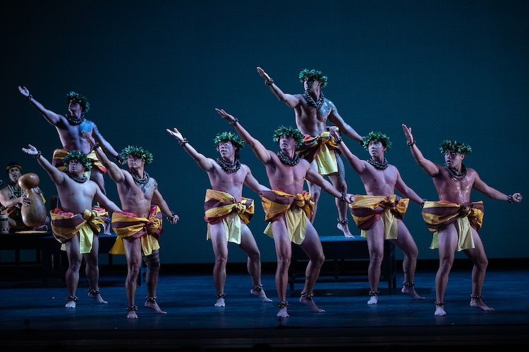 a company of male dancers from Hawaii, each raise their arms to a highh diagonal in a moment of ritual dance. the wear crowns of green leaves on their heads, necklaces that appear green, and gold red and brown wraps around the middle of their bodies... like short shorts. Behind them a musican dressed similarly sings and plays a gourd.