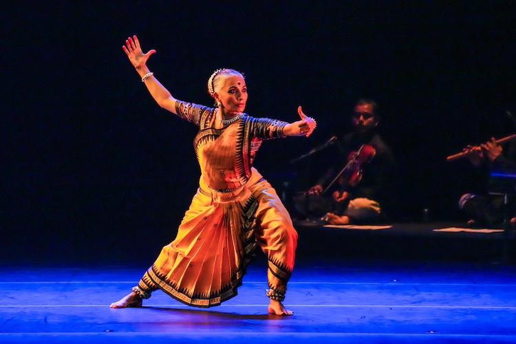 a female  Indian dancer in gold and red colors predominantly, lunges towards the audience, eyes-wide, in the middle of an exciting tale it seems... musicians play in the background