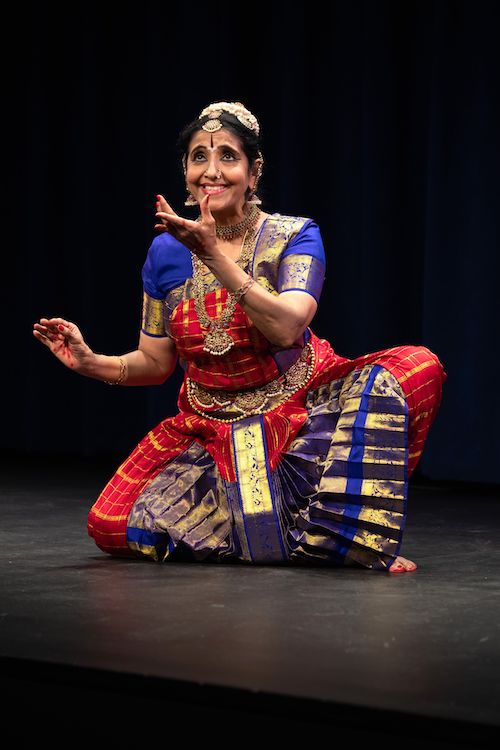 Bala Devi Chandrashekar in a bright blue and red, gold adorned traditional dance dress, raises her eyes to the sky as she gestures forward with her arm.